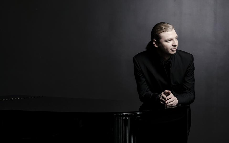 Pianist Denis Kozhukhin leaning on his instrument, looking off camera, dressed in black
