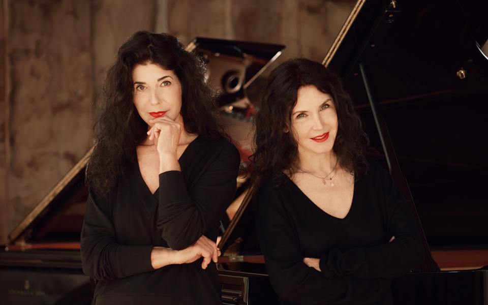 211021 Labeque sisters credit Umberto Nicoletti The pianists Katia and Marielle Labeque. The two women with long black hair and red lipstick look directly into the camera. In the background there are two Steinway Grand Pianos.