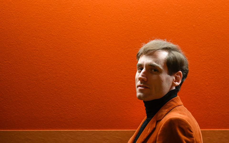 Boris Giltburg facing the side, turning his head to look to the camera. He is wearing a black turtleneck and orange jacket, stood in front of an orange wall.