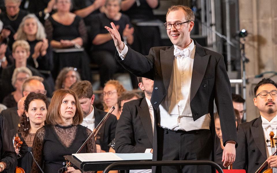 Samuel Hudson credit Dale Hodgetts for PO website Samuel Hudson reaching his arm out towards the audience and smiling, with the Philharmonia Orchestra in the background