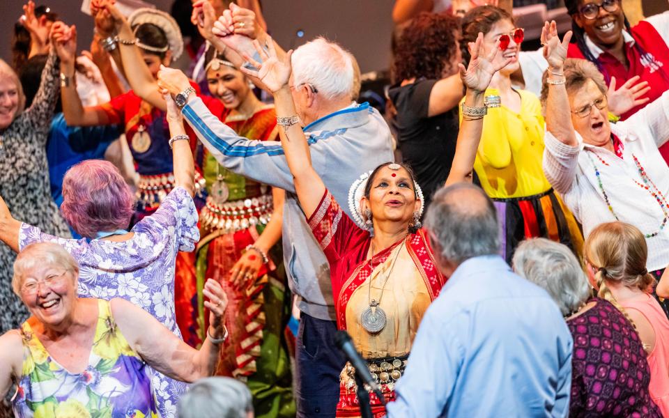 Hear and Now Celebration (001) A woman in a traditional Indian dress throws her hands up in dance, surrounded by smiling children and adults.