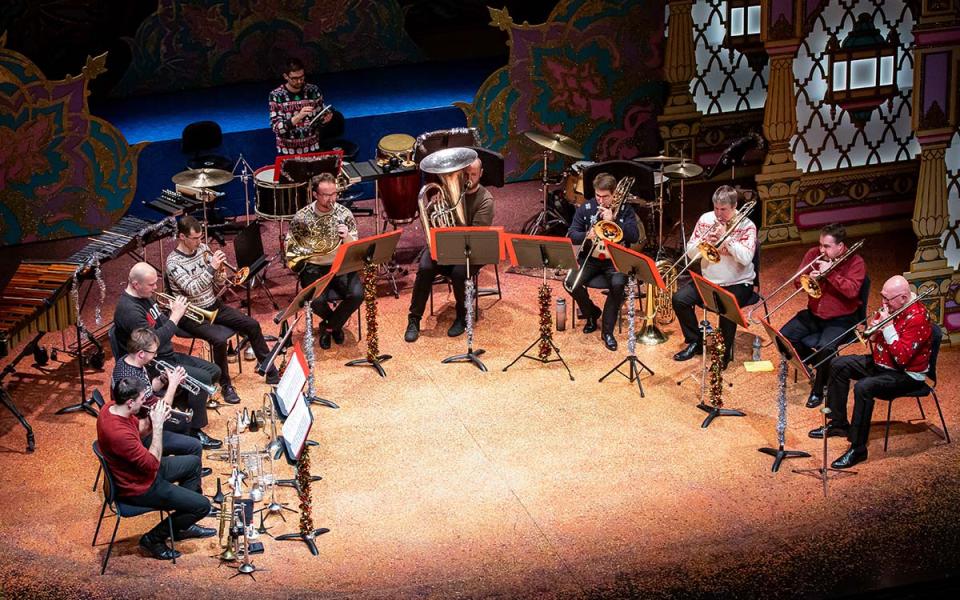 Philharmonia Brass credit Marc Gascoigne Brass players sat in a semi circle wearing casual clothes and Christmas jumpers. They are sat amongst a colourful pantomime set.
