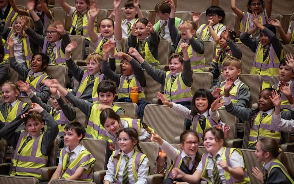 Children in an auditorium with their arms in the air, as part of the Philharmonia's Orchestra Unwrapped concert