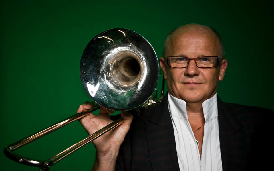 Christian Lindberg image for PO website Christian Lindberg in a white shirt and black jacket, standing in front of an emerald green background. He is holding his trombone up and looking directly into the camera