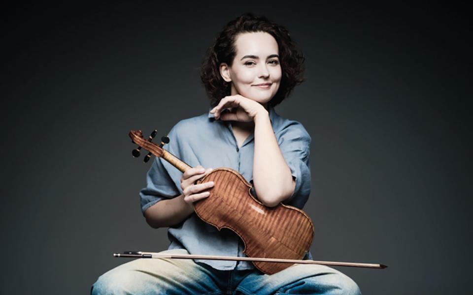 Alena Baeva sitting on Stool holding violin which is facing her. Her left arm is resting on the violin with her head resting on her hand, whilst smiling at the camera