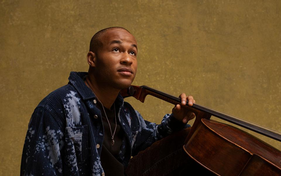 Sheku sitting on the floor with cello on his right shoulder, looking up.
