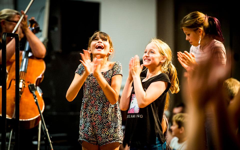 Two young girls applauding while standing at a concert, with a cellist in the background