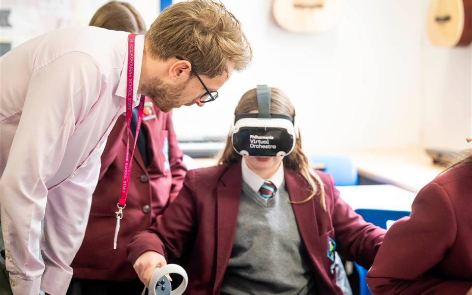 Girl in burgundy and grey school uniform, wearing a virtual reality headset and holding nunchuck, with teacher looking over to make sure she is okay.