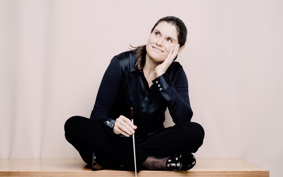 Delyana Lazarova wearing dark clothing and shoes, sat cross-legged on a bench. She is holding her baton in her hand and looking diagonally away from the camera.