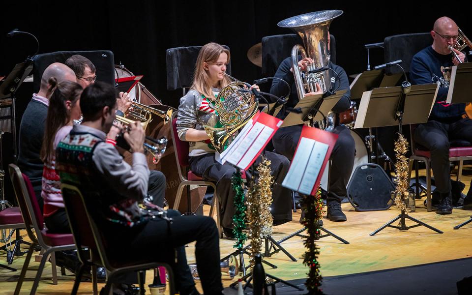 An ensemble of brass musicians on stage, wearing Christmas jumpers and with tinsel on their music stands.