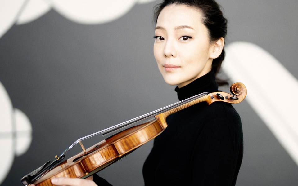 Clara-Jumi Kang resting her violin on her shoulder and looking directly into the camera. She is wearing dark clothing.