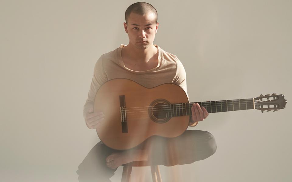 Sean Shibe sat half cross-legged on a stool, holding his guitar and staring directly into the camera. He is wearing light clothing and is against a light background.