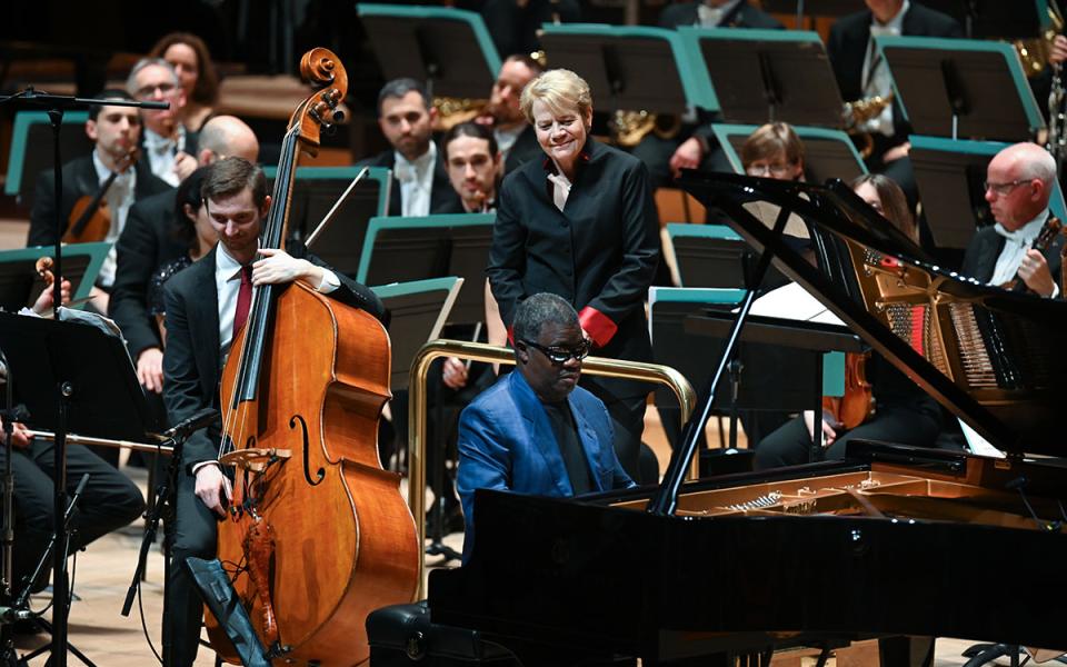 Pianist and bassist on stage in front of the orchestra, with Marin Alsop watching them and smiling