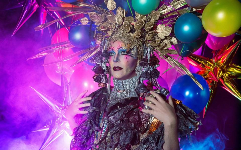 Taylor Mac in sparkling clothes, looks outwards against a colourful background of balloons
