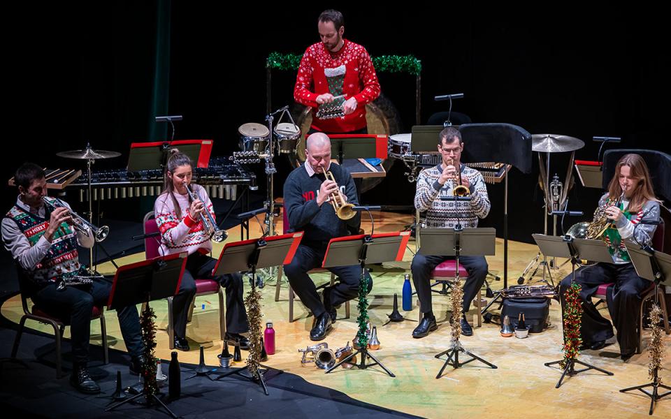 Brass musicians and one percussionist on stage, wearing Christmas jumpers with tinsel on their music stands