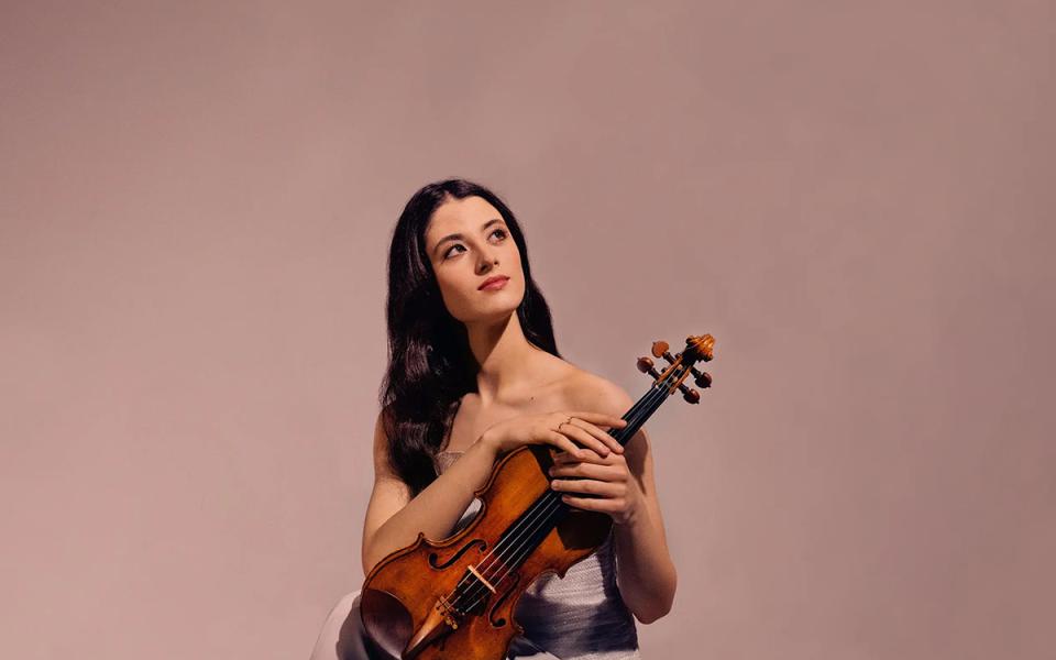 Maria Duenas against a dusty pink background holdinger her violin and looking up to the right corner