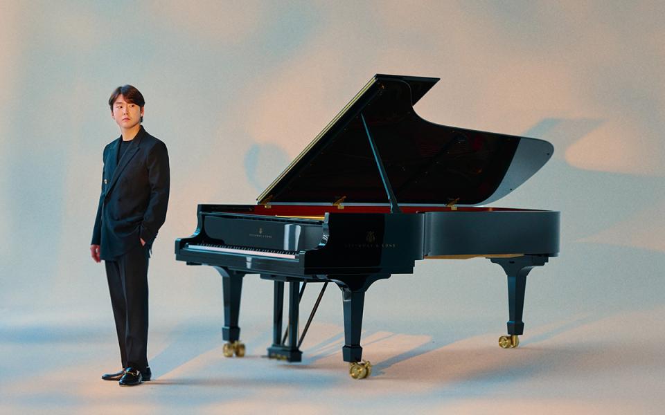 Seong-Jin Cho in light lighting in a black suit standing near a grand piano.