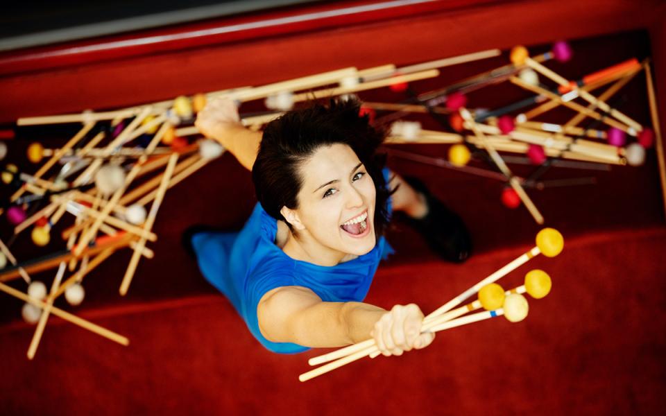 Vivi Vassileva wearing a blue dress, holding four percussion sticks up and looking up into the camera
