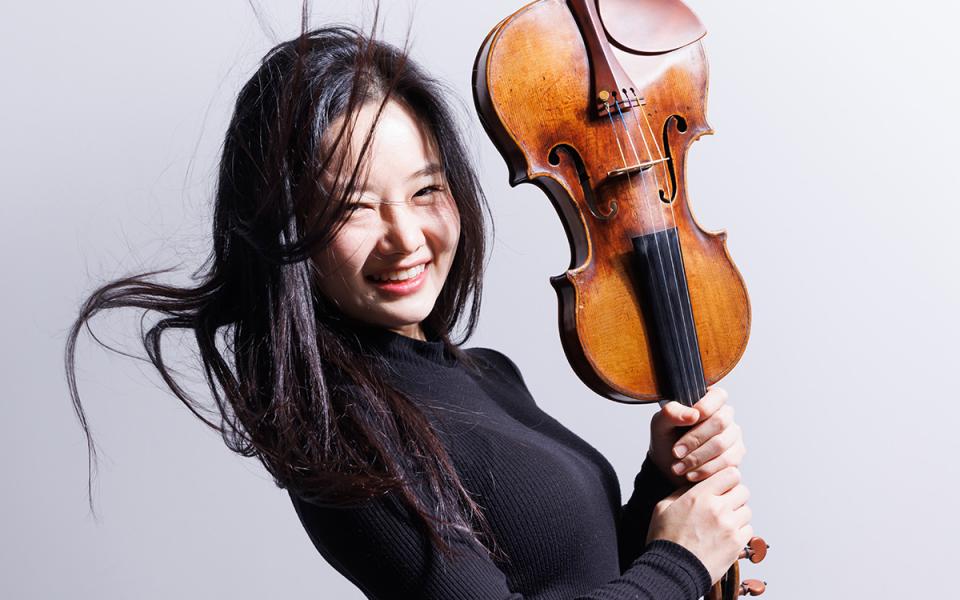 Esther Yoo in dark clothing against a white background. She is laughing and holding her violin upside down.