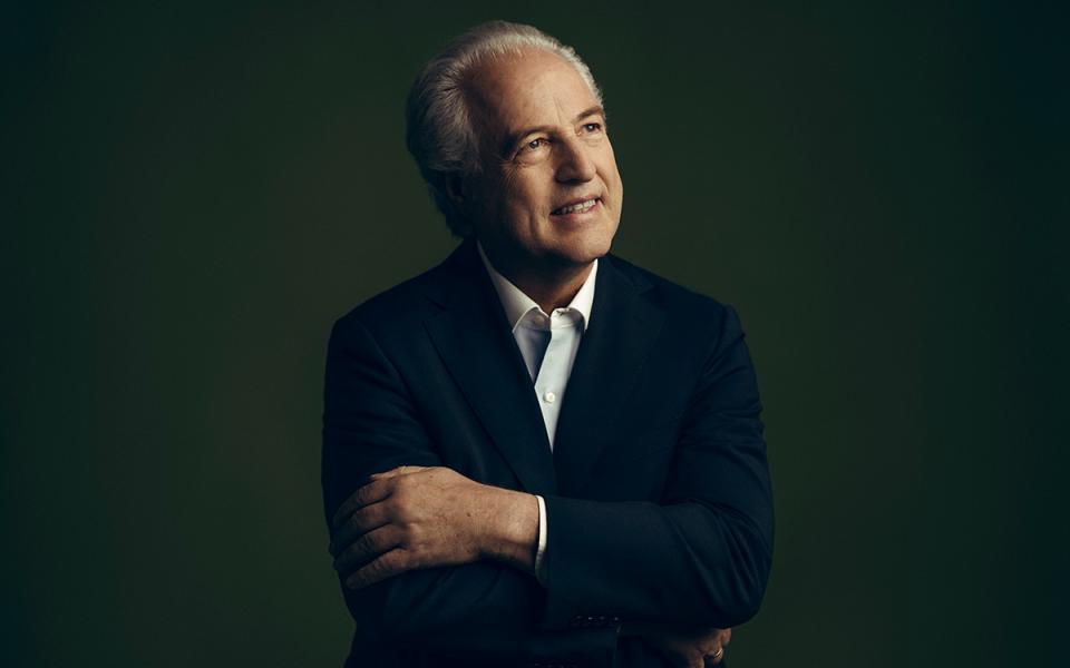 Manfred Honeck against a dark green background, crossing his arms, looking upwards into the corner and smiling